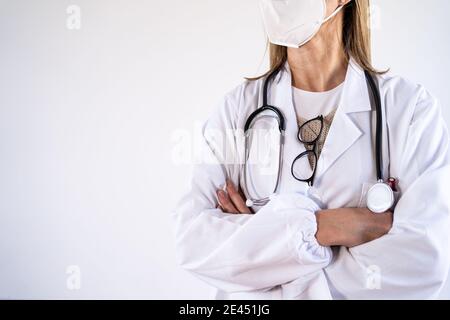 Medico femminile non riconoscibile in uniforme bianca e stetoscopio indossato maschera medica protettiva in piedi con braccia incrociate Foto Stock