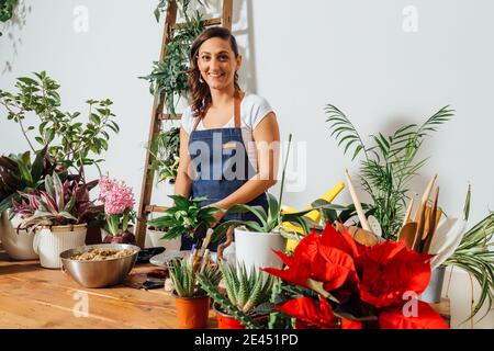 Ragazza entusiasta giardiniere in grembiule trapiantando pianta verde mentre lavora nel negozio di fiori guardando la macchina fotografica Foto Stock