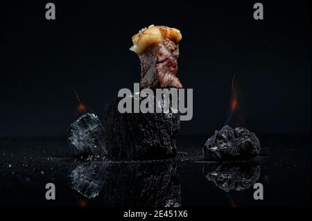 Appetitoso piccolo cubo di carne cruda con grasso posto su pezzo di carbone prima della preparazione contro sfondo nero Foto Stock