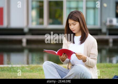 Ritratto di una donna asiatica studentessa universitaria che si apita sull'erba nel campus cercando felice e leggendo un libro nel parco. Foto Stock