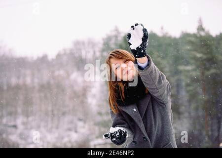 Una bella giovane ragazza con un naso rosso sorride e lancia palle di neve alla macchina fotografica sullo sfondo del paesaggio invernale. Foto Stock