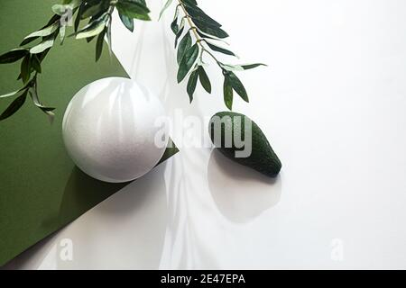 Composizione minima con avocado, forma geometrica bianca, ramo di eucalipto e ombre su sfondo bianco e verde Foto Stock