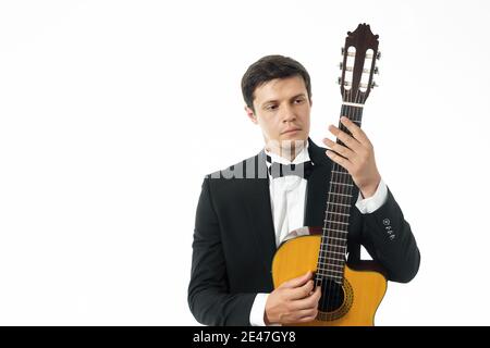 Giovane uomo in abito classico nero in posa con chitarra classica e guardando il collo di una chitarra classica. In studio su sfondo bianco Foto Stock