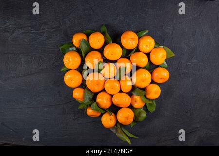 Tangerini arancioni con foglie verdi a forma di cuore su fondo di pietra nera. Cuore fatto da tangerini. Vista dall'alto. Spazio di copia. In alto Foto Stock