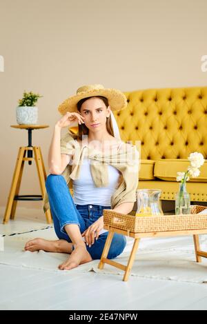 Ragazza a piedi nudi in jeans, camicia e cappello di paglia seduta sul pavimento vicino tavolo laterale con bevanda fresca Foto Stock