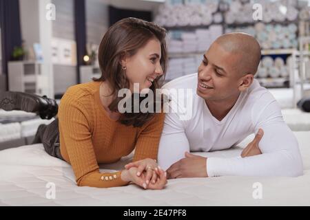 Bella coppia multirazziale sorridente l'una all'altra, sdraiata insieme su un nuovo letto in vendita al negozio di mobili Foto Stock