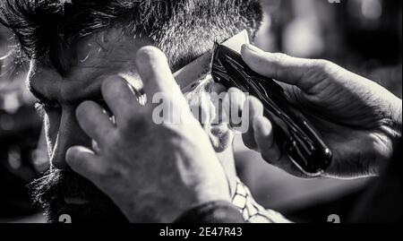 Cliente hipster ottenere haircut. Mani di barbiere con regolacapelli, primo piano. Concetto di taglio dei capelli. Uomo in visita hairstylist in barbershop. Barbiere lavori Foto Stock