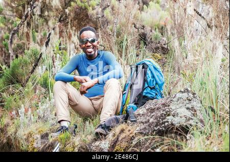 Ritratto di un giovane afroamericano Ethnicity sorridente in occhiali da sole seduti con uno zaino, riposati nella foresta e guardando il Foto Stock