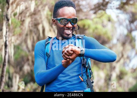 Ritratto di un allegro sorridente afroamericano Ethnicity giovane uomo controllo uno smartwatch da polso a distanza o a cuore nella foresta durante un hiki Foto Stock