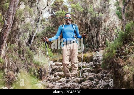 Ritratto di un giovane afroamericano Ethnicity sorridente in occhiali da sole. Ha una passeggiata con uno zaino utilizzando i bastoni da trekking a Mweka ga Foto Stock