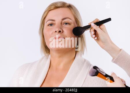 Ritratto di una donna adulta e delle mani di un maestro che si trucco con un pennello su uno sfondo bianco studio. Foto Stock