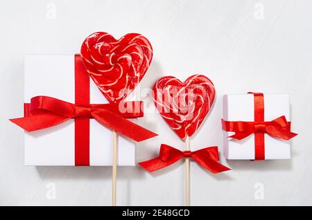 Sfondo di compleanno luminoso con amore - scatole regalo bianche con arco rosso, cuori di lecca su tavola di legno bianco, primo piano. Foto Stock