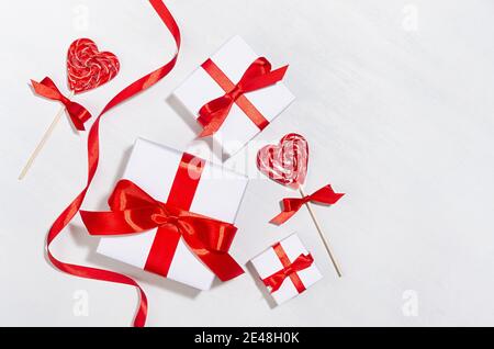 Divertimento San Valentino sfondo - scatole regalo bianche con arco rosso, cuori di lecca dolce su tavola di legno bianco, bordo. Foto Stock