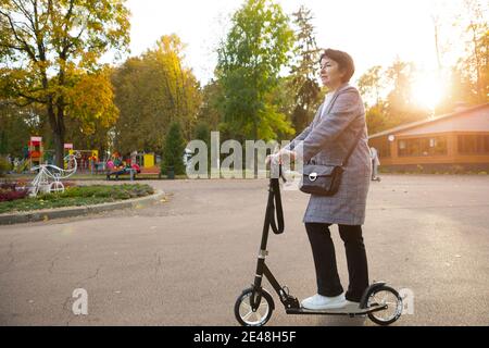 Una senora negli anni '50 su uno scooter in abiti casual gode di vita in una passeggiata nel parco. Stile di vita attivo nella mezza età, sport. Spazio di copia Foto Stock
