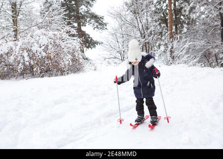 I piedi dei bambini in sci di plastica rossa con bastoni attraversano la neve da uno scivolo: Uno sport invernale, intrattenimento per tutta la famiglia all'aria aperta. Una bambina g Foto Stock