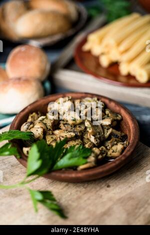 spicchio di alcuni funghi a fette cucinati con aglio e prezzemolo serviti in un piatto di terracotta, su un tavolo di legno grigio accanto ad alcuni piatti con un po 'di c Foto Stock