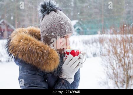 donna di mezza età che tiene una tazza di bevanda calda e sorridente sullo sfondo di una strada invernale. sparare all'aperto. Foto Stock