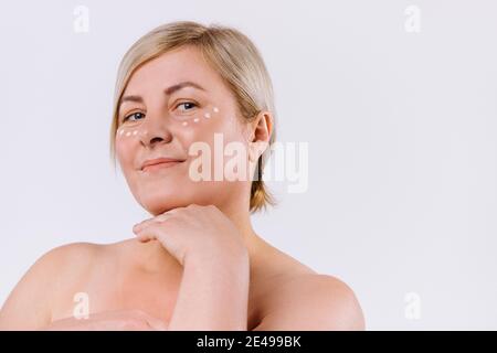 Un ritratto ingrandito di una donna bionda con gocce di una maschera idratante sotto gli occhi su sfondo bianco. Foto Stock