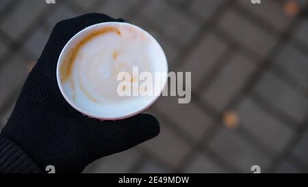 Un bicchiere di cappuccino in mano a mans, un giovane beve il cappuccino mentre tiene una tazza di caffè in guanti invernali. Foto Stock