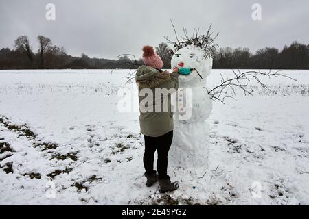 Donna in abiti invernali facendo un simpatico pupazzo di neve aggiungendo medico maschera facciale Foto Stock