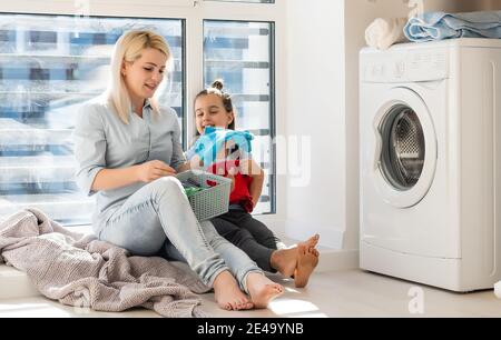 madre di famiglia e bambina piccola aiutante in lavanderia vicino alla lavatrice e ai vestiti sporchi Foto Stock