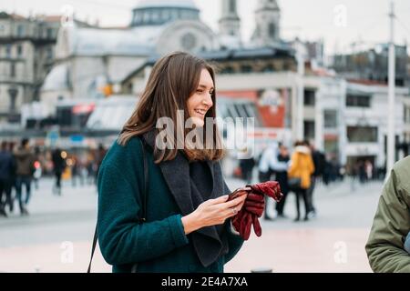 TURCHIA, ISTANBUL, 14 DICEMBRE 2018: Bella ragazza in un cappotto con uno smartphone su Piazza Taksim sorride felicemente. Foto Stock