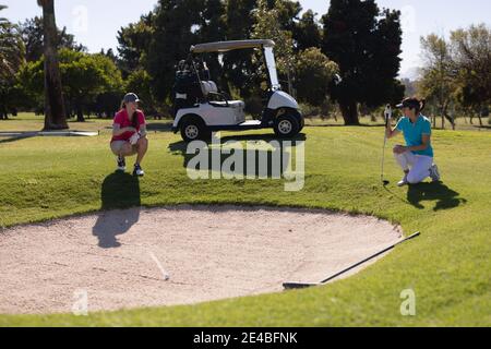 Due donne caucasiche giocare a golf squatting giù accanto bunker che parla Foto Stock