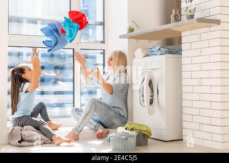 madre di famiglia e bambina piccola aiutante in lavanderia vicino alla lavatrice e ai vestiti sporchi Foto Stock