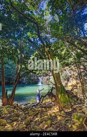 Una donna (MR) circondata da alberi si trova in uno stagno con una cascata in una giungla tropicale sull'isola di Maui, Hawaii. Foto Stock