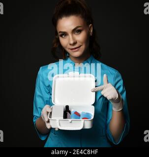Giovane donna sorridente medico infermiera in uniforme blu e protettivo guanti in lattice che tengono il kit di pronto soccorso a mani e sguardo alla telecamera Foto Stock