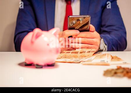 banca dei piggy, monete e cinquanta euro in primo piano. Uomo d'affari che usa lo smartphone mobile in background Foto Stock