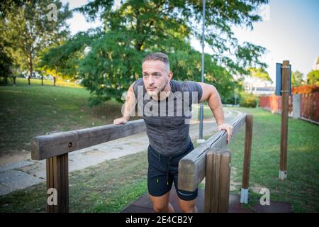 Uomo europeo in pantaloncini neri e t-shirt grigia che si tuffa su barre parallele. Allenamento all'aperto in un parco Foto Stock