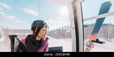 Winter sport Asian woman sitting in ski lift going up the hill at ski resort going skiing down the slopes. Banner panoramic of skier wearing helmet Foto Stock