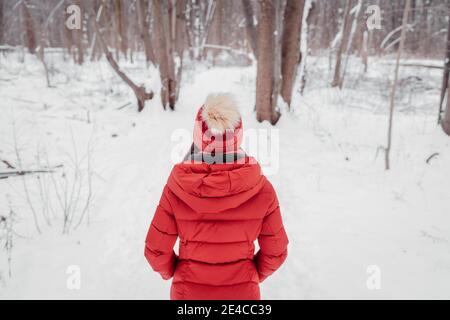 Donna invernale che cammina nel parco dei boschi innevati. percorso forestale all'aperto. Vista da dietro della donna in cappotto rosso e berretto Foto Stock