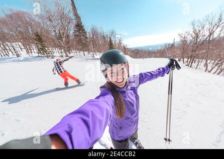 Sport invernali divertimento - felice sciatore asiatico prendendo selfie con amico snowboarder in background. Stazione sciistica vacanze piste Foto Stock