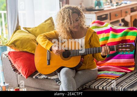 Bella donna di mezza età suona la chitarra a casa - musica lezione e cantante seduti sul divano - persone che giocano strumentale e godendo le canzoni e le attività di svago all'interno Foto Stock