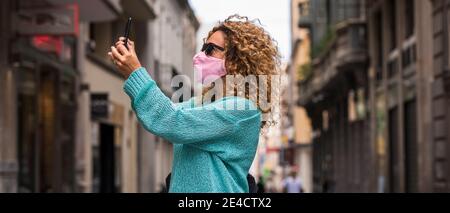 Donna fare video telefono chiamata indossare maschera di protezione medica per coronavirus covid-19 emergenza sanitaria in città - persone a piedi e godere della città protetta dal virus del contagio Foto Stock