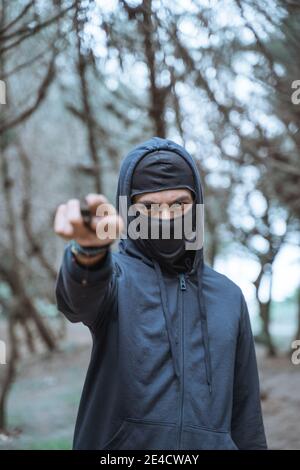 uomo in una maschera con un coltello che indossa abiti neri sullo sfondo di una foresta Foto Stock