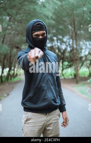 cattivo in una maschera con un coltello indossando una felpa nera con cappuccio puntare il coltello verso la fotocamera quando si trova sul strada Foto Stock