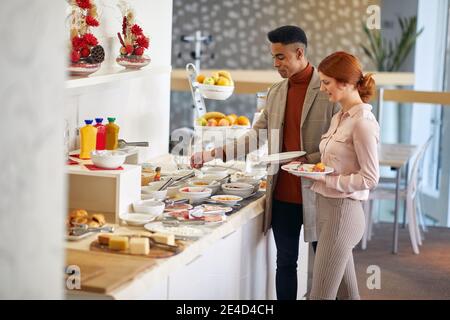 uomo afro-americano che parla con la donna caucasica durante la pausa pranzo, scegliendo il cibo da un buffet Foto Stock