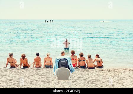 Rodi, Grecia - Luglio 11 2017: Uomo maturo in piedi in acqua e gruppo di donne che si siedono sulla spiaggia di sabbia il giorno estivo in resort Foto Stock