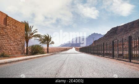 Lunga strada dritta con colline desertiche a distanza, muro di mattoni e recinzione di ferro sui lati. Scenario all'ingresso dell'area protetta di Wadi Rum della Giordania Foto Stock