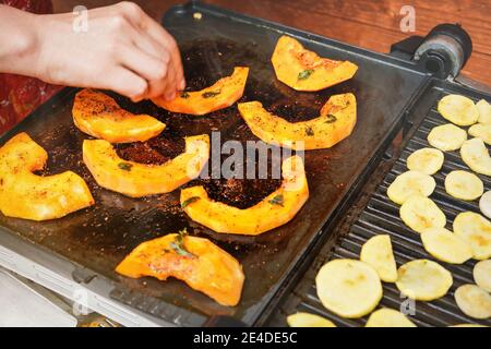 Pezzi di zucca arancione brillante alla griglia elettrica, mano della donna sfocata che muove pezzi di verdure sopra il piatto caldo Foto Stock