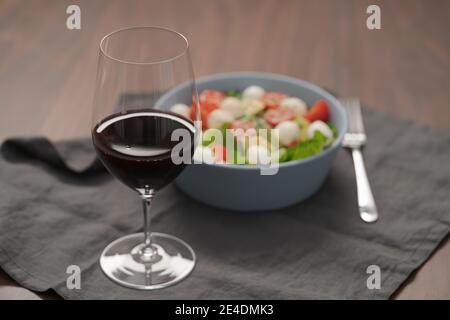 Bicchiere di vino rosso con insalata con romaine, pomodori e mozzarella in ciotola blu, a fuoco basso Foto Stock