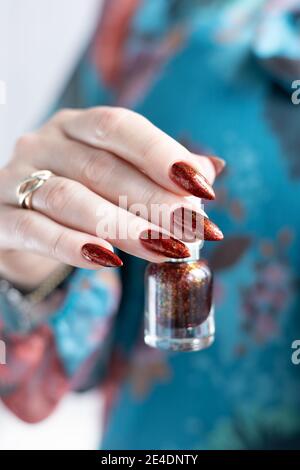 Mano femminile con unghie lunghe e marrone dorato, manicure contiene una bottiglia di smalto per unghie Foto Stock