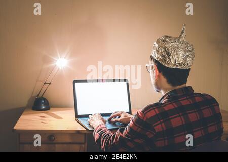 Un giovane uomo con cappuccio in alluminio è seduto nel seminterrato scuro di fronte a un computer portatile. Concetto di teoria della cospirazione Foto Stock