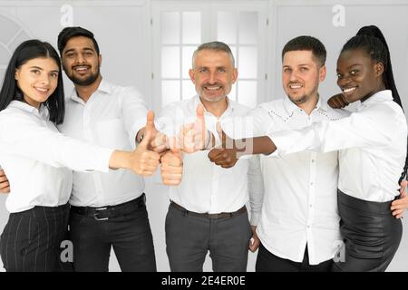 Gruppo di felici e sorridenti impiegati internazionali in camicie bianche mostrare i pollici verso l'alto e guardare la fotocamera Foto Stock