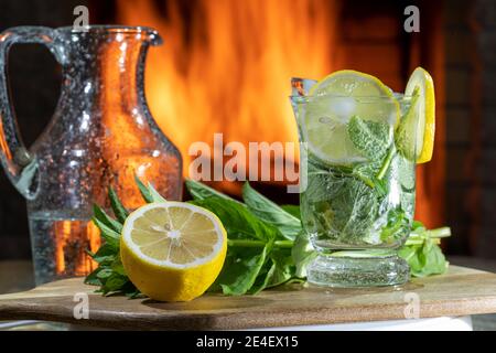 Limonata con acqua minerale, limone, menta e ghiaccio su un tavolo di legno prima di accogliente camino. Bevanda analcolica. Foto Stock