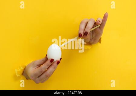 Tema astratto di buona Pasqua. Mani che dipingono un uovo di pasqua bianco mentre si rompono attraverso i ritagli di uno sfondo di cartone giallo Foto Stock