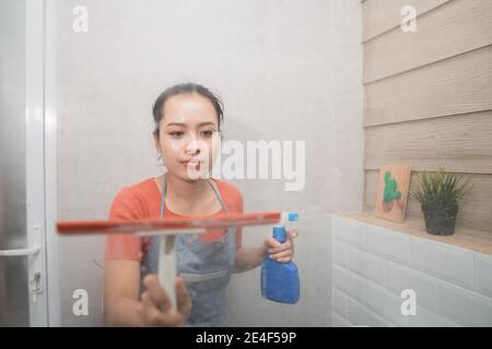 Donna asiatica salviette con un tergicristallo e tiene un spruzzatore di bottiglia mentre pulisce il vetro del wc nel bagno Foto Stock